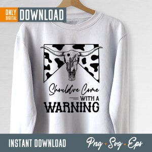 Op de afbeelding: Een wit sweatshirt met een zwart-wit grafisch ontwerp van een koeien schedel met hoorns en een cactus. De tekst "Should've Come With A Warning" staat onder de schedel.