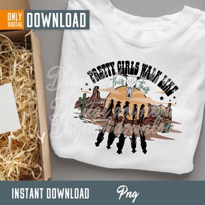 May include: A white t-shirt with a graphic design of a sunset scene with cacti and a cow skull. The text "Pretty Girls Walk Like This" is written in a green and black font around the graphic.