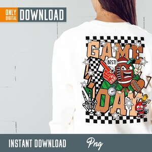 Op de afbeelding: Een wit T-shirt met een grafisch ontwerp van een voetbalpersonage met de woorden "Game Day" op een geruite achtergrond. Het ontwerp bevat sterren, een voetbal, een fluitje, een klok en een trofee.