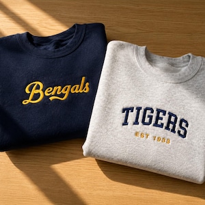 Peut inclure: Un sweat-shirt bleu marine à col rond avec le mot Bengals brodé en écriture jaune et un sweat-shirt gris clair à col rond avec le mot TIGERS brodé en lettres capitales bleu marine au-dessus du texte EST 1933 brodé en jaune.