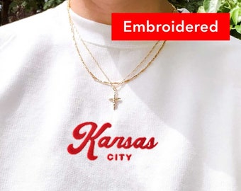 May include: White sweatshirt with red embroidered text that says "Kansas City" and a gold chain necklace with a cross pendant. The text is labeled "Embroidered".