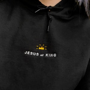 以下が含まれることがあります： 黒いフード付きのスウェットシャツに、白い刺繍で「JESUS is KING」と書いてあり、その上に小さな黄色の王冠が刺繍されています。
