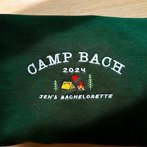 May include: Green sweatshirt with white embroidery that says "Camp Bach 2024" with a red heart and a small embroidered image of a yellow tent, a campfire, and two pine trees. The text "Jen's Bachelorette" is also embroidered below the main text.