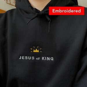 以下が含まれることがあります： 黒いフード付きのスウェットシャツで、金の刺繍の王冠と「JESUS is KING」の文字が前面に刺繍されています。