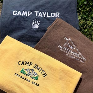 Op de afbeelding: Drie t-shirts in geel, bruin en donkergrijs. Het gele t-shirt heeft geborduurde tekst "CAMP SMITH" met een berg- en boomafbeelding en "COLORADO 2026". Het bruine t-shirt heeft een wimpelafbeelding met "CAMP WILLIAMS" en "YELLOWSTONE 2027". Het donkergrijze t-shirt heeft "CAMP TAYLOR" en een berenpootafdruk.