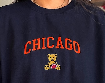 Könnte beinhalten: Marineblaues Sweatshirt mit dem Wort "CHICAGO" in orangefarbener Stickerei, über einem gelben Teddybär, der einen Football hält. Das Sweatshirt hat einen Rundhalsausschnitt.