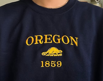 Può includere: Una felpa blu navy con testo ricamato in giallo che recita "OREGON" e una grafica di castoro con l'anno "1859" sotto.