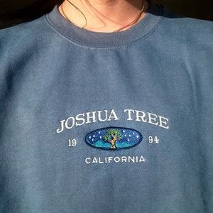 May include: A blue sweatshirt with a white embroidered design that says "Joshua Tree 19 84 California" with a stylized Joshua Tree inside an oval.