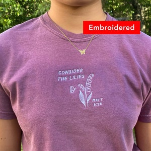 May include: A purple t-shirt with an embroidered design featuring a lily flower and the text "Consider the lilies" and "Matt 6:28".