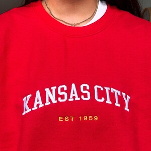 Puede incluir: Una sudadera roja con el texto blanco "KANSAS CITY" en arco. Debajo, el texto dorado dice "EST 1959". La sudadera es de color rojo liso.
