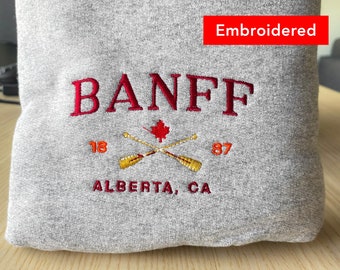 Puede incluir: Sudadera gris con texto bordado en rojo que dice "BANFF 1887 ALBERTA, CA" y una hoja de arce roja con dos remos naranjas cruzados.