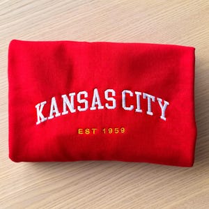 Puede incluir: Sudadera roja doblada con texto bordado blanco "KANSAS CITY" y el a&ntilde;o "EST 1959" en dorado. La sudadera est&aacute; hecha de un material suave y est&aacute; doblada cuidadosamente.