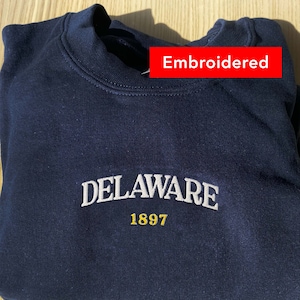 Peut inclure: Un sweat-shirt bleu marine avec le texte "DELAWARE 1897" brodé en blanc et or sur le devant.
