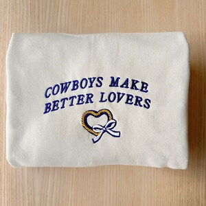 May include: White sweatshirt with blue embroidered text that reads "Cowboys Make Better Lovers" and a yellow heart with a blue ribbon.