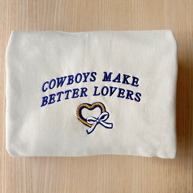 May include: White sweatshirt with blue embroidered text that reads "Cowboys Make Better Lovers" and a yellow heart with a blue ribbon.