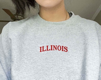 May include: A light gray sweatshirt with the word "ILLINOIS" embroidered in red across the chest. The sweatshirt has a crew neck and a relaxed fit, perfect for casual wear.