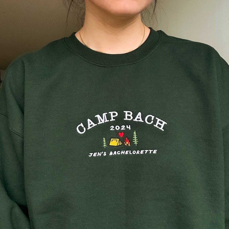 May include: A dark green sweatshirt with white embroidery that reads "Camp Bach 2024" with a small red heart and a yellow tent graphic. The text "Jen's Bachelorette" is also embroidered in white.