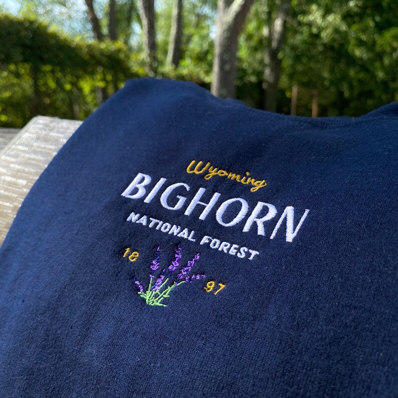 May include: A navy blue t-shirt with white embroidered text that reads "Wyoming BIGHORN NATIONAL FOREST 18 97" and a purple and green embroidered flower design.