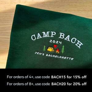 May include: Green sweatshirt with embroidered text "CAMP BACH 2024" and a small camping scene with a tent, fire, and heart. The text "JEN'S BACHELORETTE" is also embroidered below the scene. The text "For orders of 4+, use code BACH15 for 15% off" and "For orders of 8+, use code BACH20 for 20% off" is printed below the image.