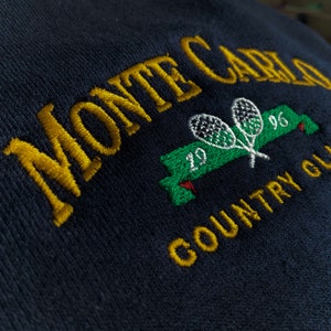May include: Embroidered navy blue patch with gold lettering that reads "Monte Carlo Country Club" and a green banner with two white tennis rackets and the number "1996" in gold.
