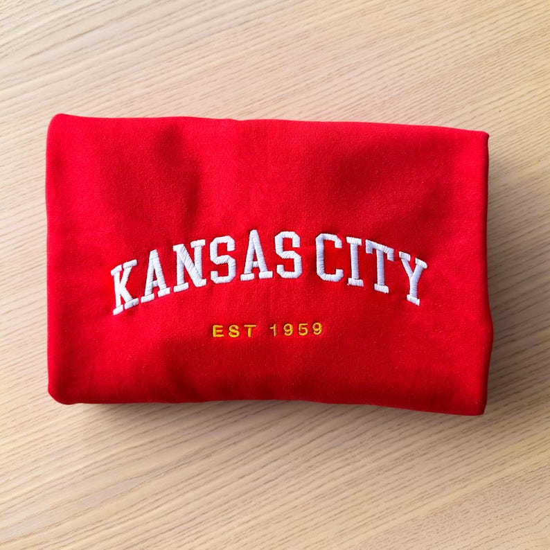 Puede incluir: Sudadera roja doblada con texto bordado blanco "KANSAS CITY" y el a&ntilde;o "EST 1959" en dorado. La sudadera est&aacute; hecha de un material suave y est&aacute; doblada cuidadosamente.