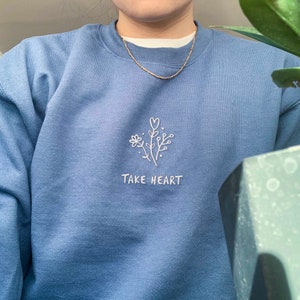 May include: A blue sweatshirt with a white embroidered design of a flower with a heart and the text "TAKE HEART".