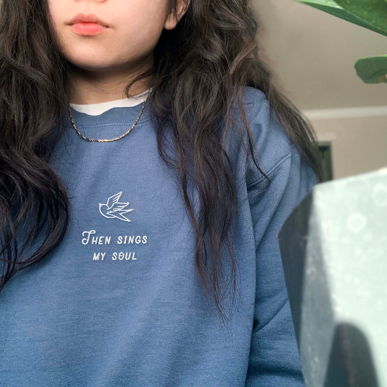 Puede incluir: Una sudadera azul con un p&aacute;jaro bordado en blanco y el texto "Then Sings My Soul".