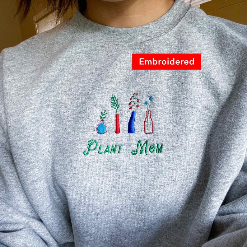 May include: Gray sweatshirt with embroidered green text that says "PLANT MOM" and a design of five embroidered plants in vases. The text "Embroidered" is written in red above the design.