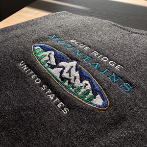 Op de afbeelding: Geborduurde patch met een blauwe en witte berg sc&egrave;ne in een ovale vorm. De tekst "Blue Ridge Mountains" staat boven de sc&egrave;ne en "United States" eronder. De patch is op een donkergrijze stof.