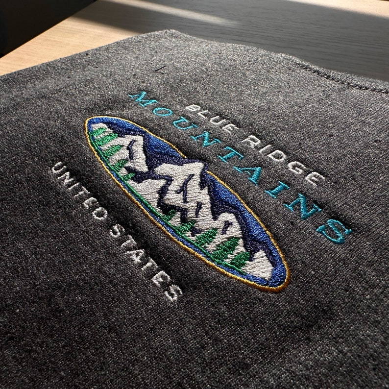 Op de afbeelding: Geborduurde patch met een blauwe en witte berg sc&egrave;ne in een ovale vorm. De tekst "Blue Ridge Mountains" staat boven de sc&egrave;ne en "United States" eronder. De patch is op een donkergrijze stof.