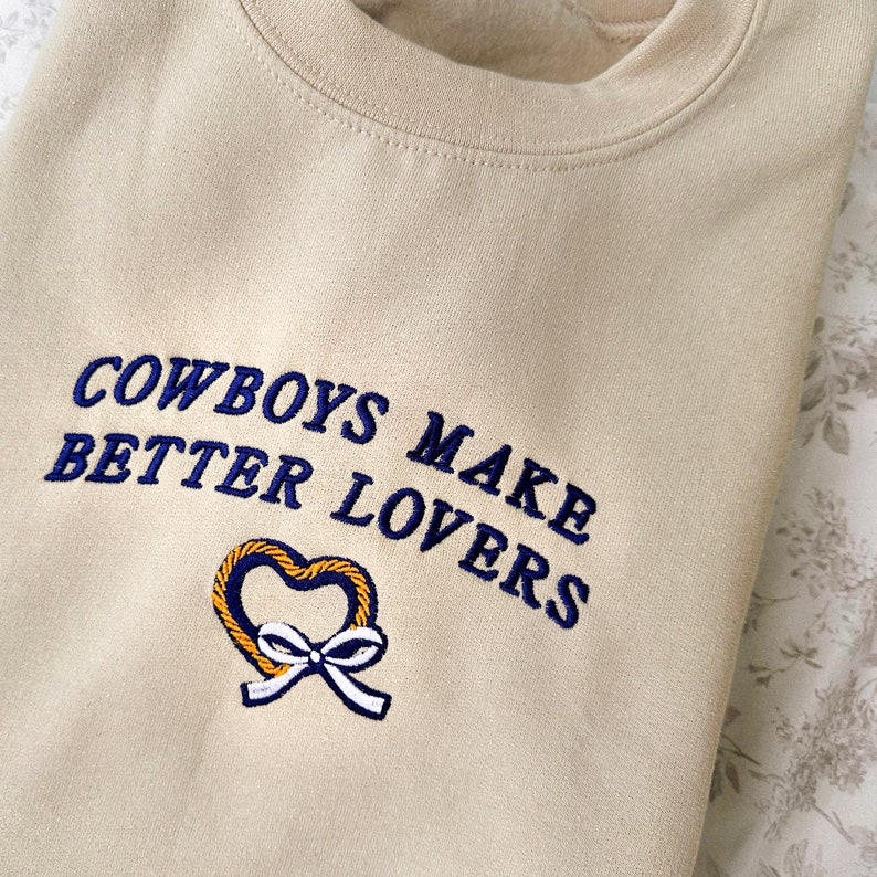 May include: A beige sweatshirt with the embroidered text "COWBOYS MAKE BETTER LOVERS" in navy blue. A yellow heart with a blue bow is embroidered below the text.