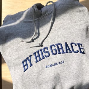 May include: Grey hooded sweatshirt with the text "By His Grace" in blue embroidered lettering and the text "Romans 3:24" in smaller lettering below.