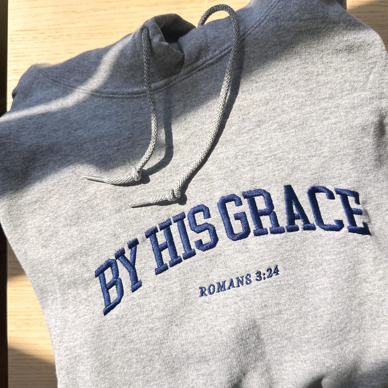 May include: Grey hooded sweatshirt with the text "By His Grace" in blue embroidered lettering and the text "Romans 3:24" in smaller lettering below.