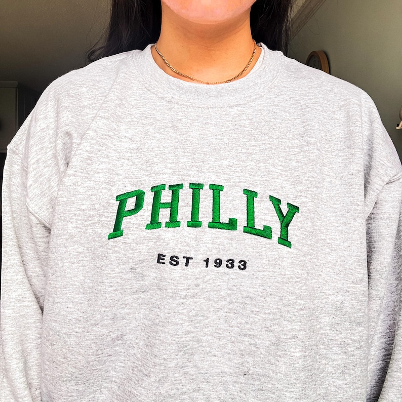 May include: A heather gray sweatshirt with the word "PHILLY" embroidered in green, arched across the chest. Below the word "PHILLY" is the text "EST 1933" in black. The sweatshirt is a casual style.