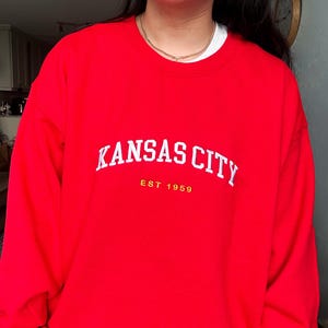 Puede incluir: Una sudadera roja con la inscripci&oacute;n blanca "KANSAS CITY" en forma de arco. Debajo, en amarillo, est&aacute; "EST 1959". La sudadera es de color rojo liso y vibrante.
