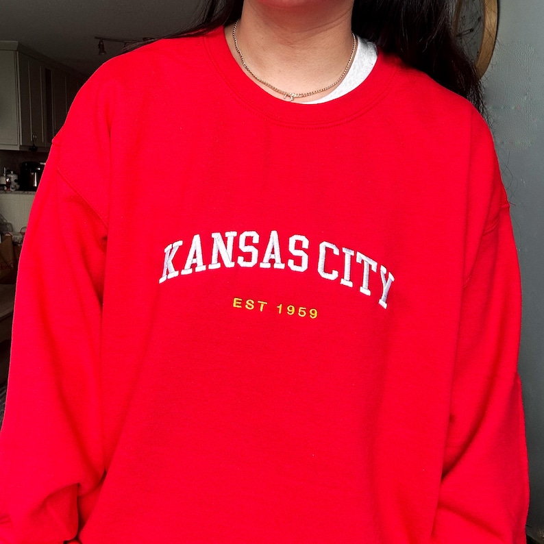 Puede incluir: Una sudadera roja con la inscripci&oacute;n blanca "KANSAS CITY" en forma de arco. Debajo, en amarillo, est&aacute; "EST 1959". La sudadera es de color rojo liso y vibrante.