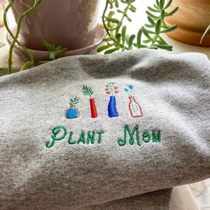 May include: Gray sweatshirt with green embroidery that reads "PLANT MOM" and features five stylized plants in vases. The vases are red, blue, and white.