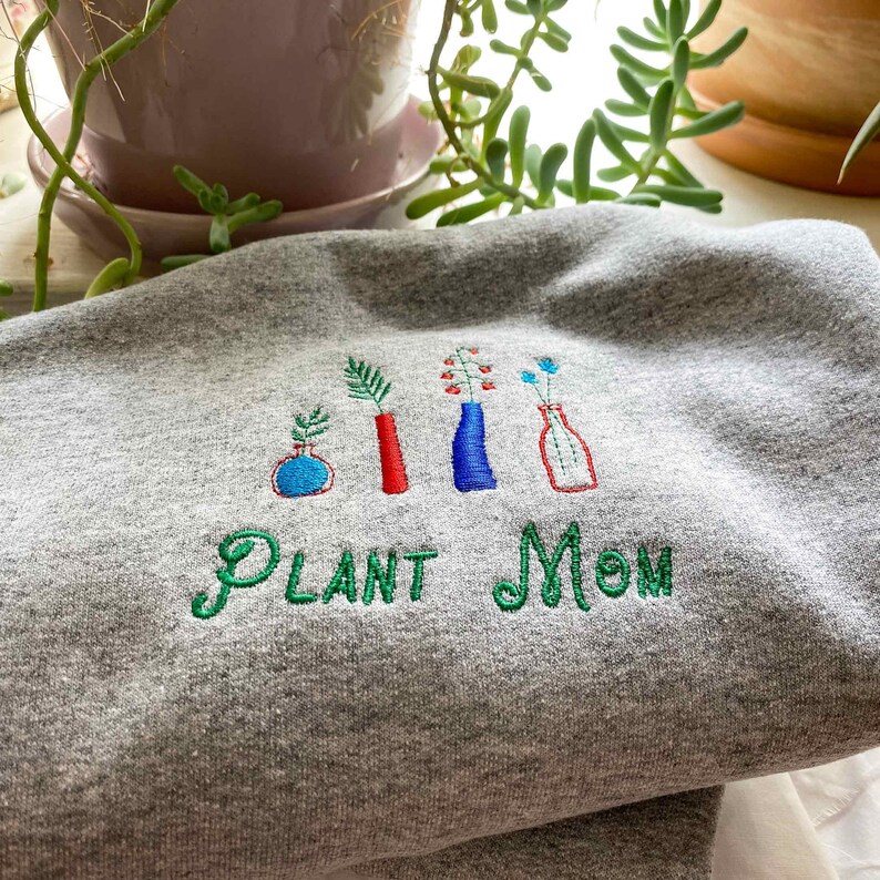 May include: Gray sweatshirt with green embroidery that reads "PLANT MOM" and features five stylized plants in vases. The vases are red, blue, and white.