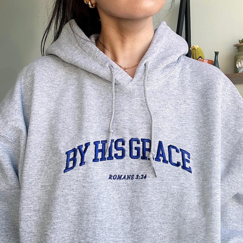May include: Grey hooded sweatshirt with the text "BY HIS GRACE" in blue embroidered lettering and "ROMANS 3:24" in smaller lettering below.