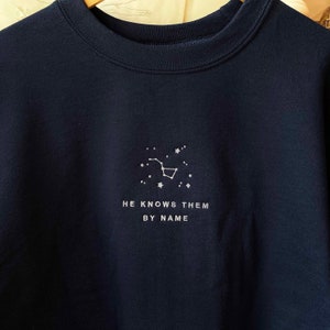 Puede incluir: Una sudadera azul marino con una constelaci&oacute;n bordada en blanco y el texto "HE KNOWS THEM BY NAME".