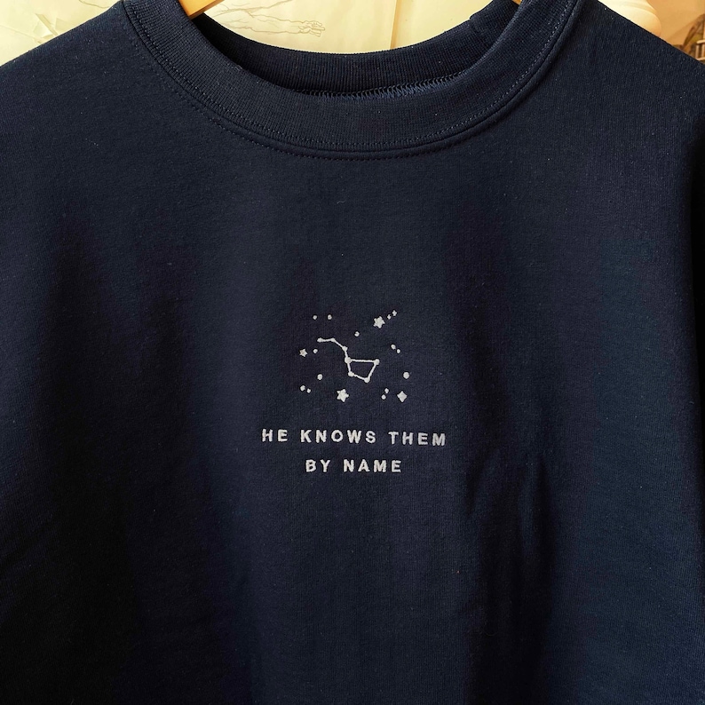 Puede incluir: Una sudadera azul marino con una constelaci&oacute;n bordada en blanco y el texto "HE KNOWS THEM BY NAME".
