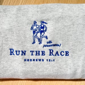 Puede incluir: Una sudadera gris con bordados azules. El texto dice "Run the Race" y "Hebreos 12:1" con un dibujo de dos corredores. La frase "with perseverance!" también está bordada en azul.