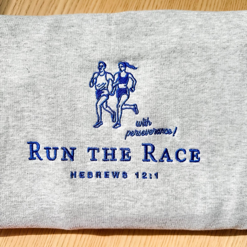May include: A gray sweatshirt with blue embroidered text and graphics. The text reads "Run the Race" and "Hebrews 12:1" with a graphic of two runners. The phrase "with perseverance!" is also embroidered.