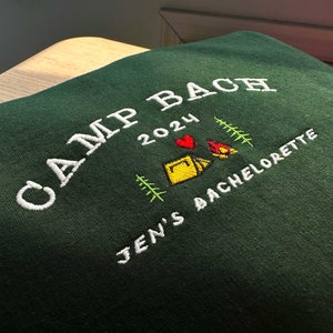 May include: Green sweatshirt with white embroidery that says "Camp Bach 2024" with a heart, tent, and campfire graphic. Below the text is "Jen's Bachelorette"