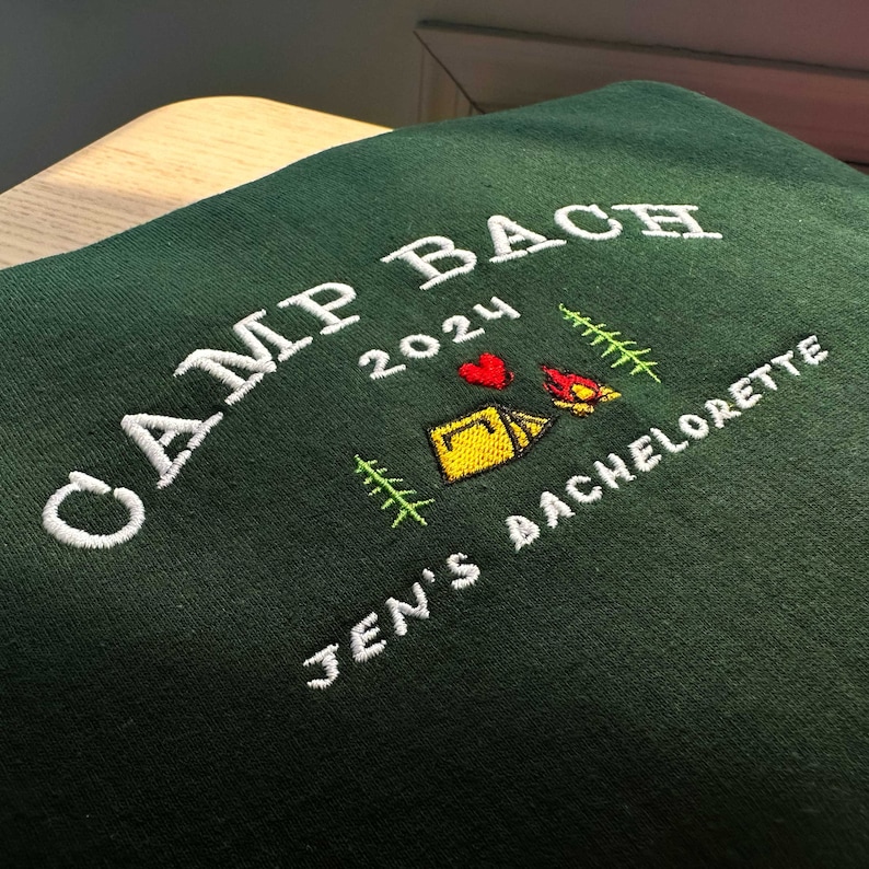 May include: Green sweatshirt with white embroidery that says "Camp Bach 2024" with a heart, tent, and campfire graphic. Below the text is "Jen's Bachelorette"