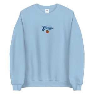 K&ouml;nnte beinhalten: Hellblauer Pullover mit dem Wort "Georgia" in blauer Schrift und einem Pfirsich-Motiv gestickt.