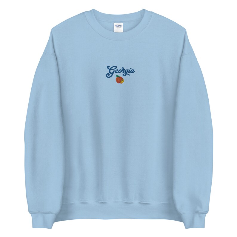 K&ouml;nnte beinhalten: Hellblauer Pullover mit dem Wort "Georgia" in blauer Schrift und einem Pfirsich-Motiv gestickt.