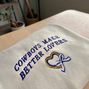 May include: A cream-colored sweatshirt with blue embroidery that reads "Cowboys Make Better Lovers" and a gold heart with a white ribbon.