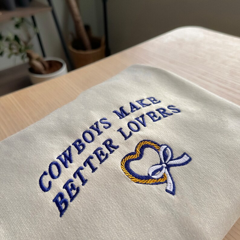 May include: A cream-colored sweatshirt with blue embroidery that reads "Cowboys Make Better Lovers" and a gold heart with a white ribbon.