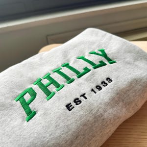 May include: A light gray sweatshirt with the word "PHILLY" embroidered in green block letters. Below the word is "EST 1933" in black. The sweatshirt is folded on a wooden surface.